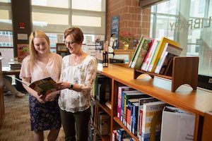 Two women standing inside of a library looking at a book